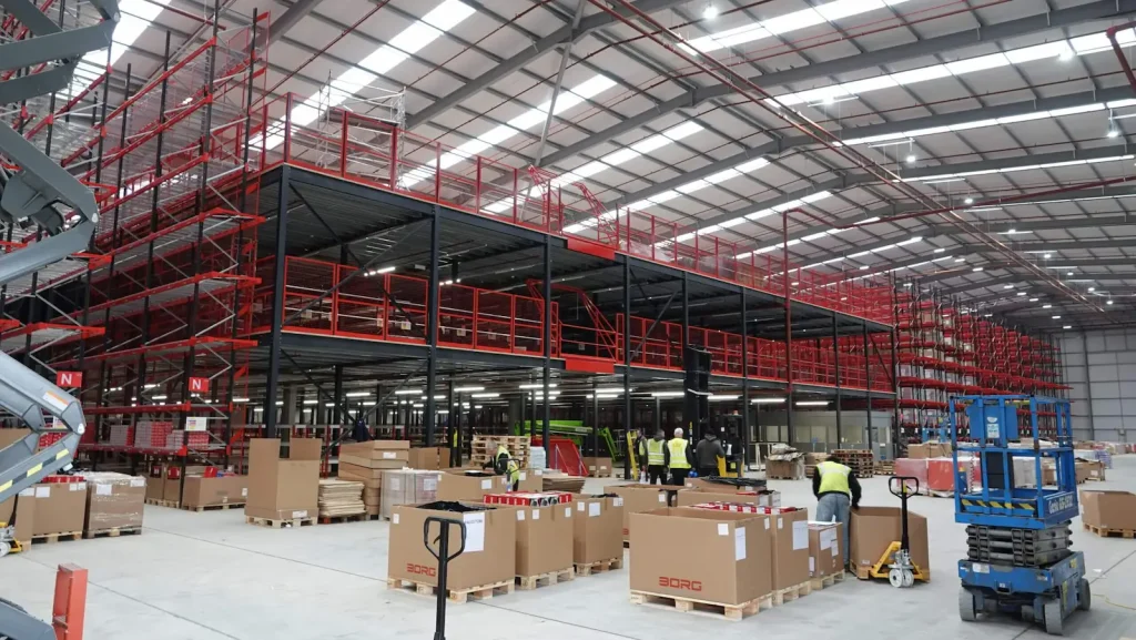 The inside of a warehouse with several pallets and boxes on display with workers wearing hi vis jackets.