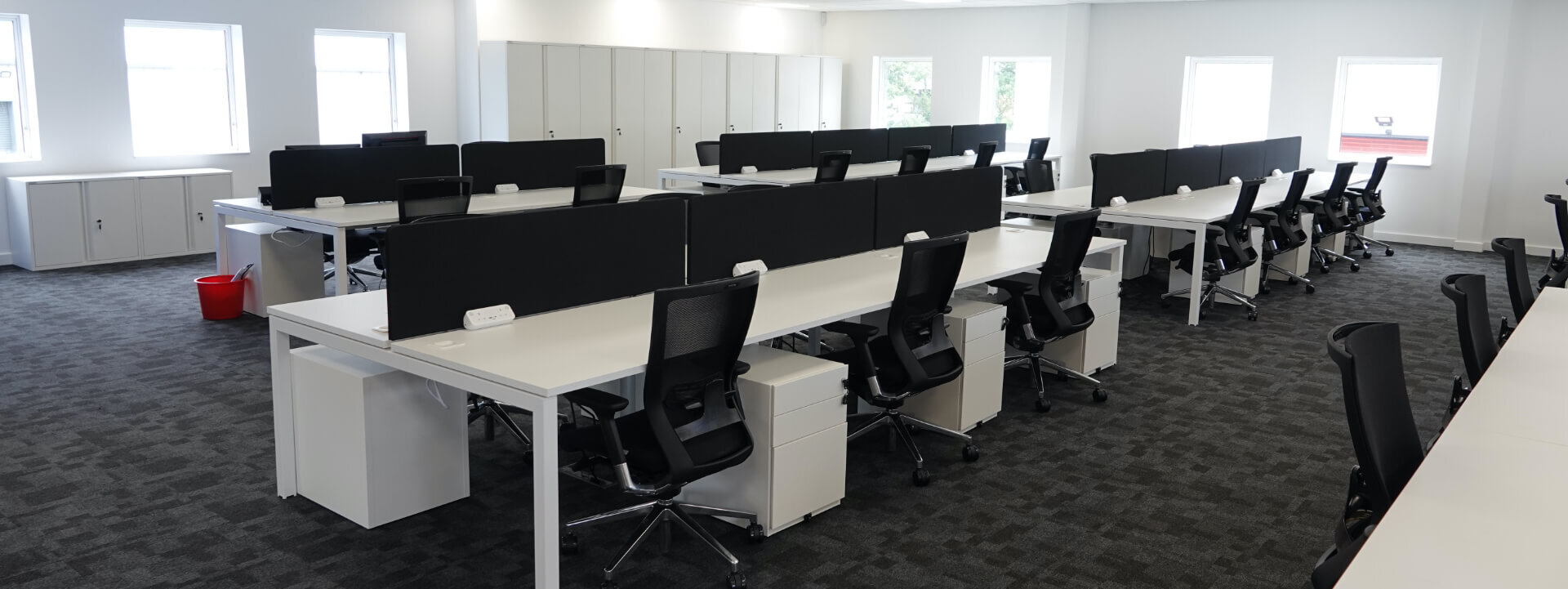 The inside of an office, there are rows of white desks with black monitors and desk chairs on a grey pattern carpeted floor.